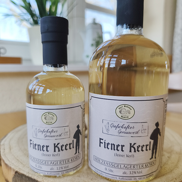 Zwei Flaschen Fiener Keerl holzfassgelagerter Korn vom Grafschafter Genusswerk in den Größen 0,2 Liter und 0,5 Liter auf einer Holzscheibe.